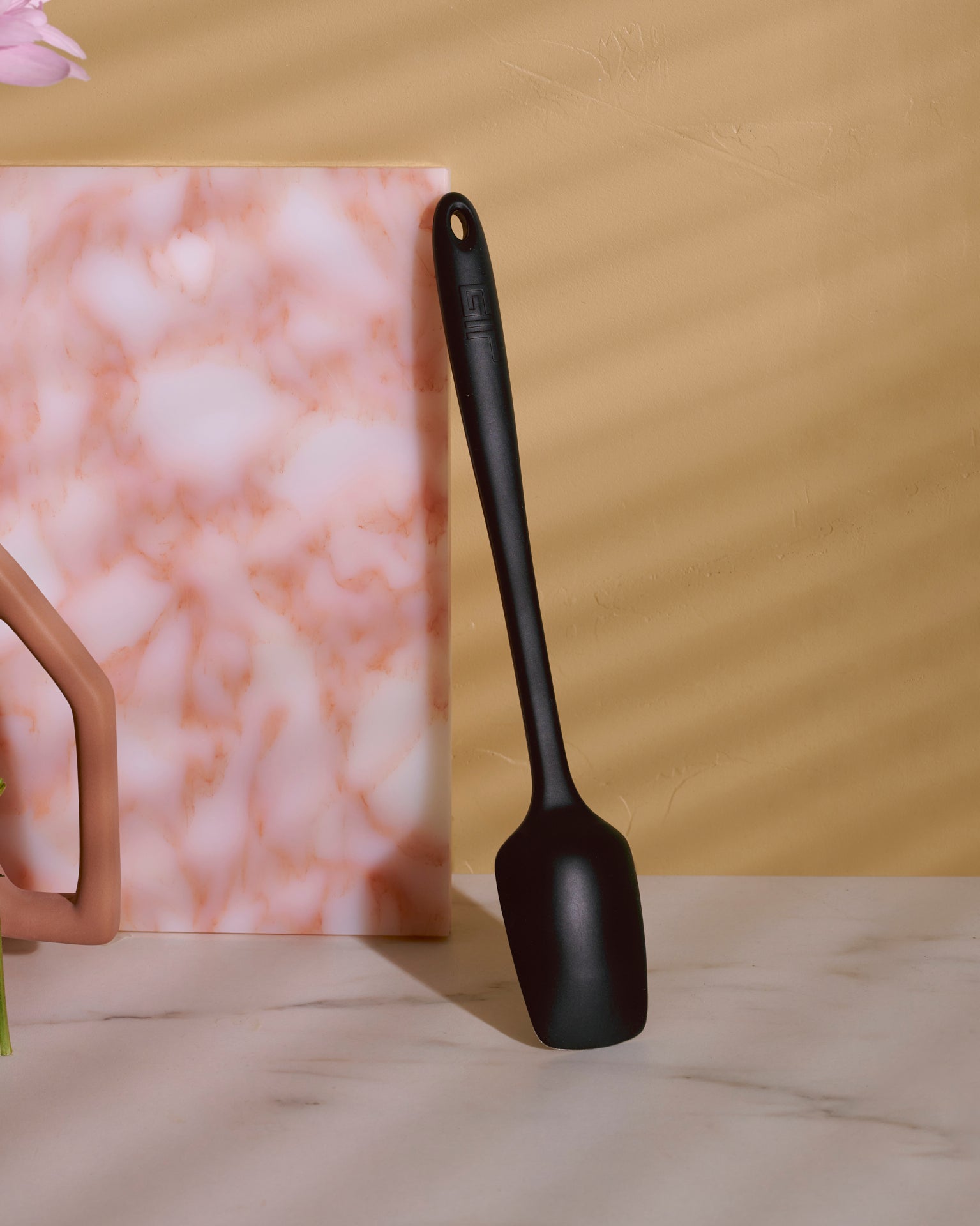 The Black Ultimate Spoonula resting upright on a white marble counter top. 